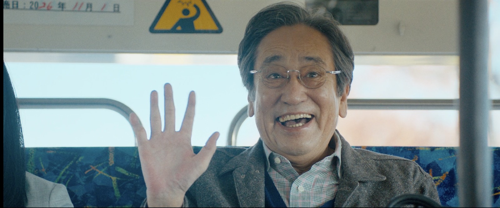 バスの座席で手を挙げて笑う豊。